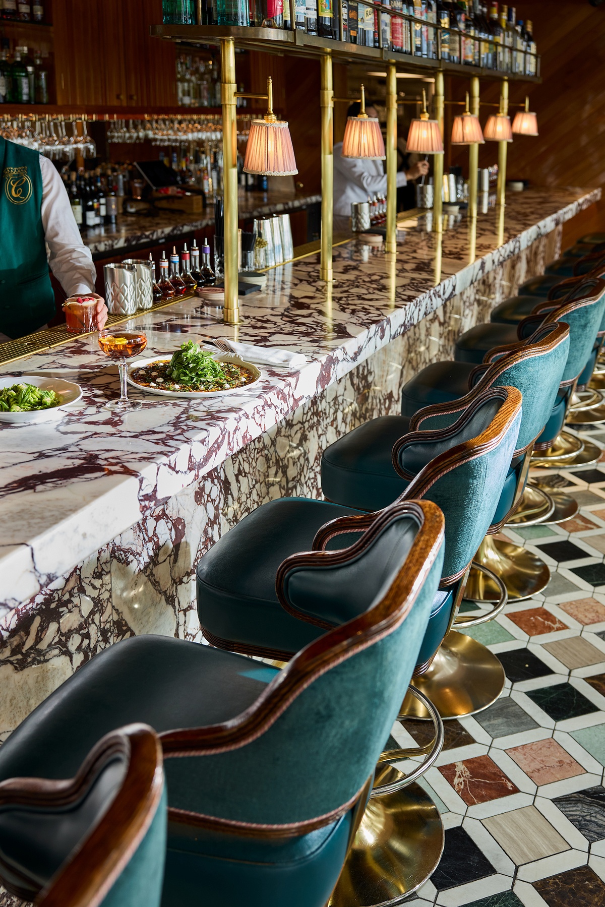 Food and drinks sit on an elegant black and white marbled bar with green velvet seating on a colorful tile floor.