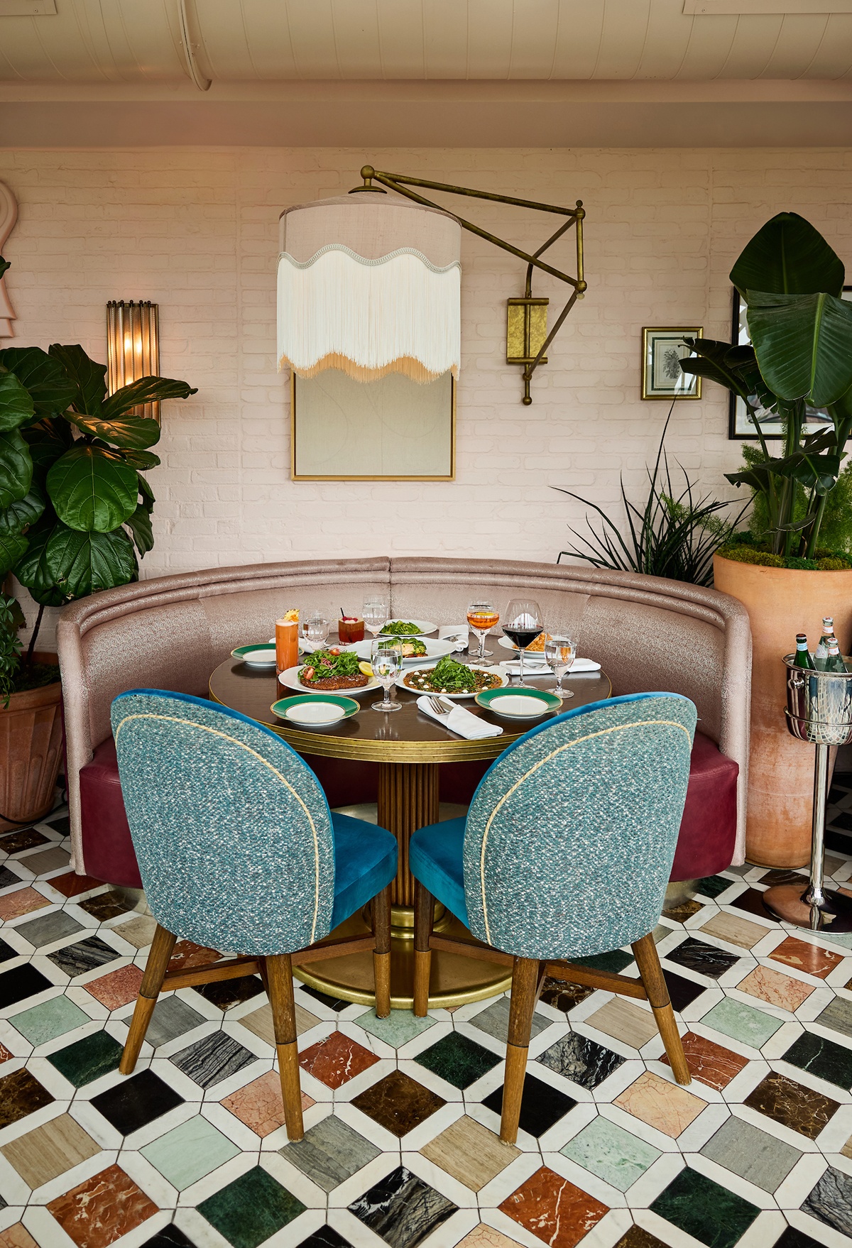 A table with a round pink booth and teal upholstered chairs on a colorful tiled floor is set with elegant plates of Italian food.