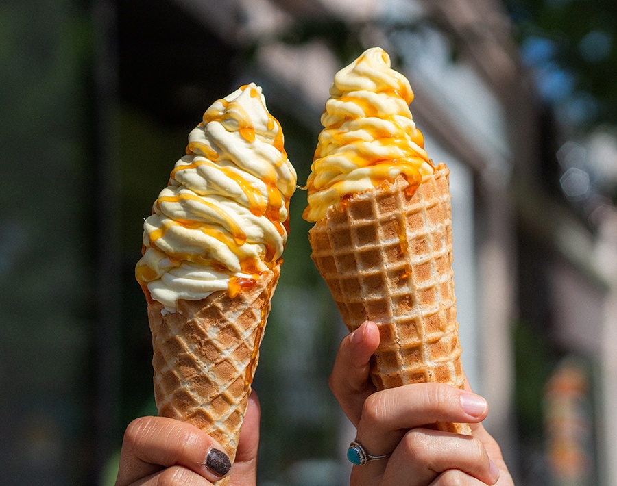 Two hands hold up cones with swirls of orange and yellow soft serve ice cream.