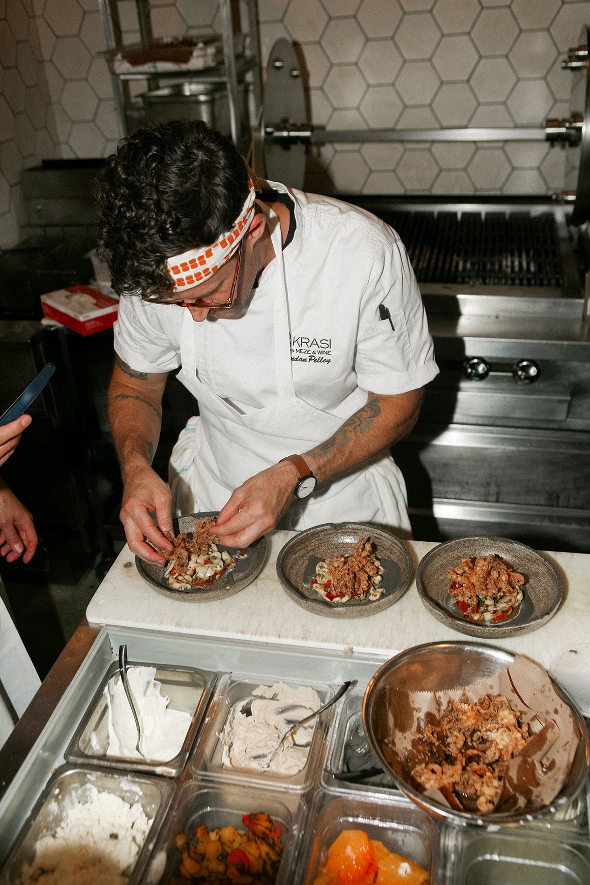 A chef in a white apron plates dishes in a restaurant kitchen with a meat spit visible in the background.