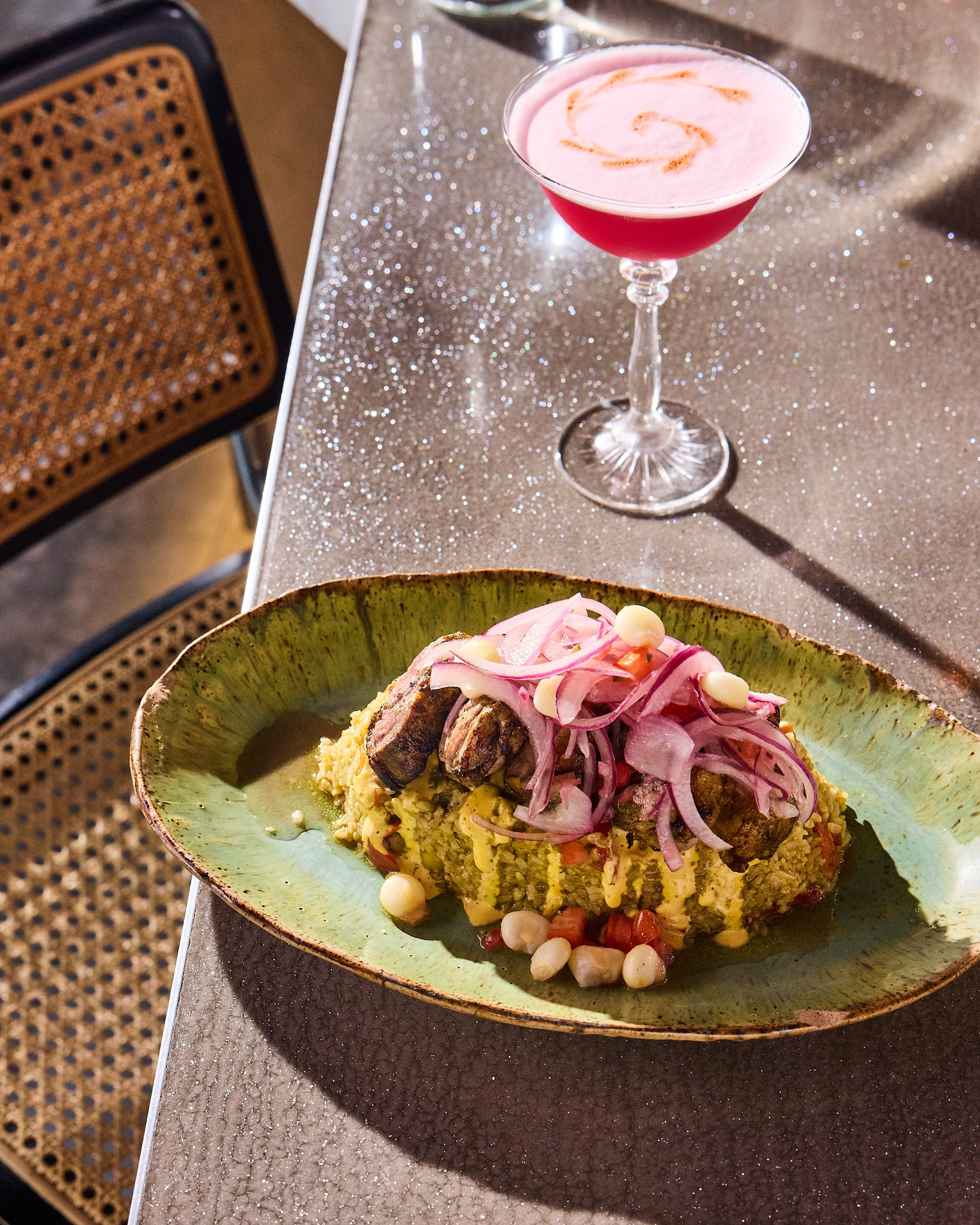 A hefty portion of rice is topped with sliced meat and red onions, sitting on a glittery silver bar with a pink cocktail alongside.