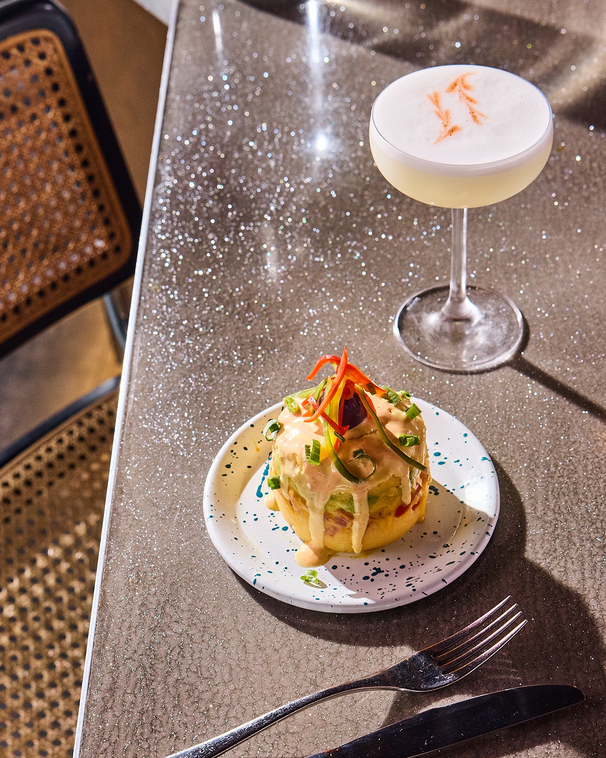 A Peruvian dish that stacks avocado atop potato sits on a glittery silver bar next to a cocktail.