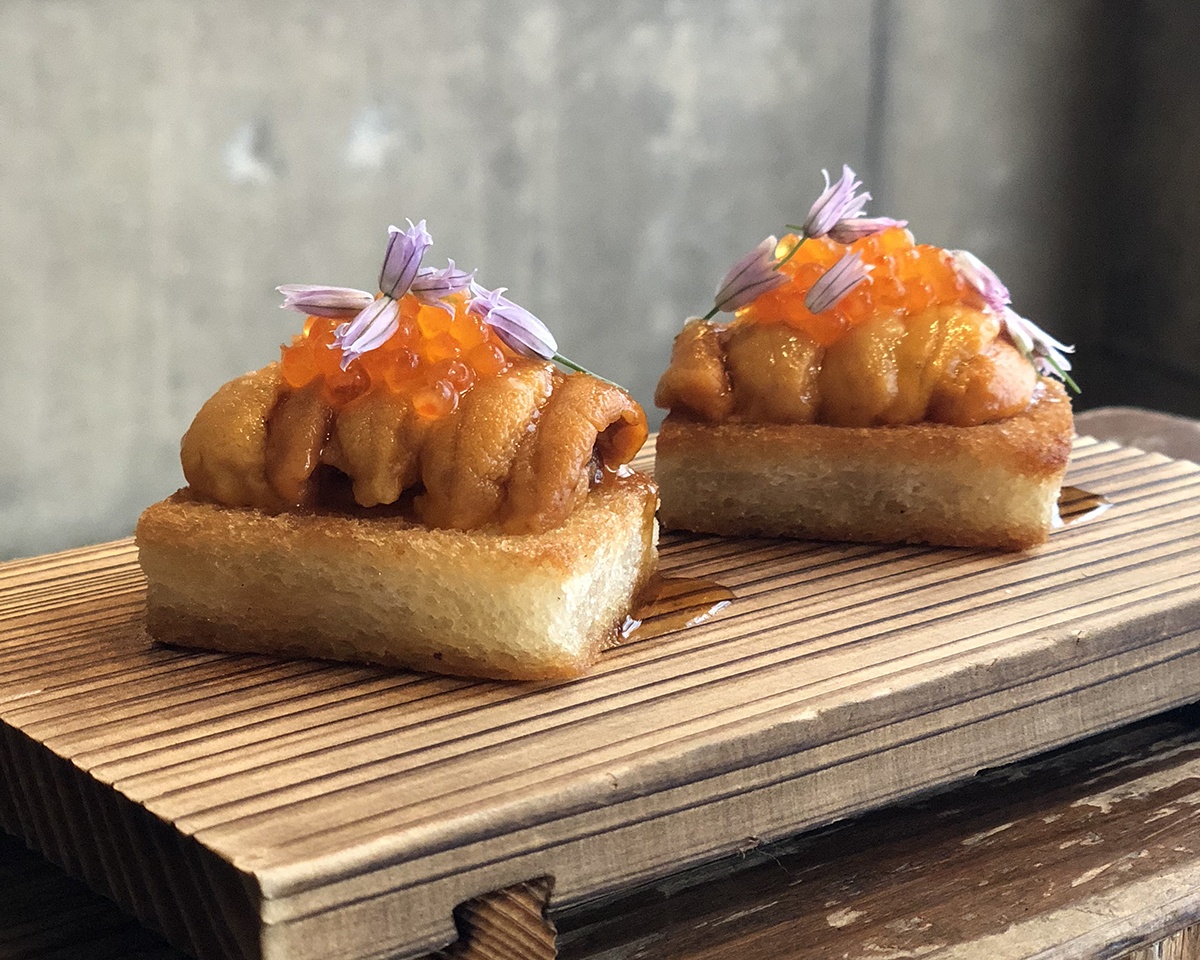 Two small rectangles of toasted bread are topped with uni, ikura, and tiny purple flowers.