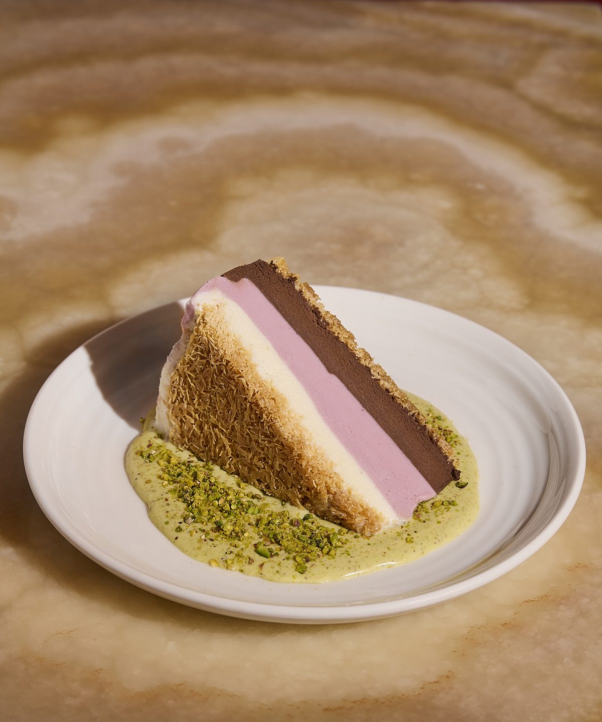 A wedge of spumoni-like ice cream cake features stripes of white, pink, and brown ice cream, sandwiched between shredded phyllo and sitting in a pool of pistachio sauce.