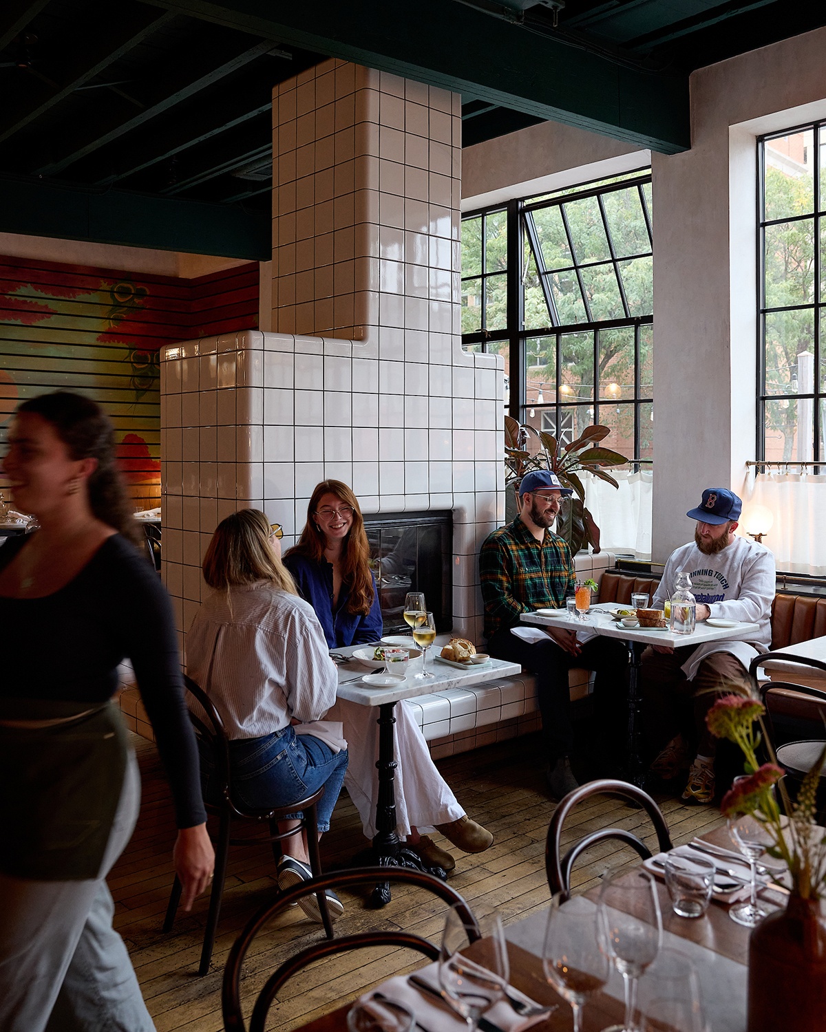 Diners sit in a restaurant with a white tiled fireplace, big windows, and a colorful mural along one wall.