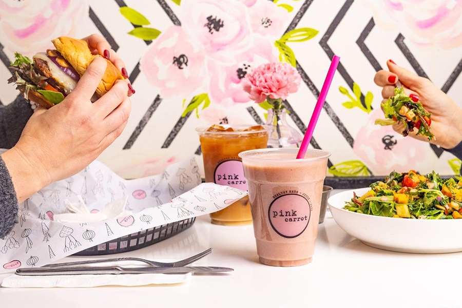 The hands of two people are visible at a white table, eating a sandwich and a salad. The wallpaper has pink flowers, and there's a pink smoothie on the table.