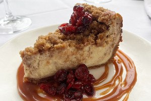 A slice of cheesecake has a coffee cake-style crumbly topping, with pickled cranberry garnish. It sits in a swirl of caramel.