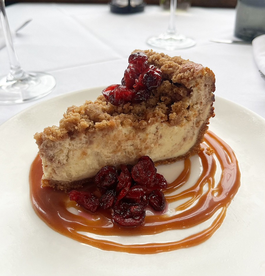 A slice of cheesecake has a coffee cake-style crumbly topping, with pickled cranberry garnish. It sits in a swirl of caramel.