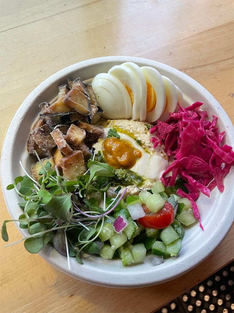 Overhead view of a bowl of fried eggplant, hummus, pickled onions, hard boiled eggs, cucumbers, and more.