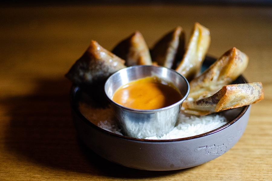 Little fried dough packets are lined up atop dry rice in a bowl, with a small metal cup of thick orange dipping sauce.