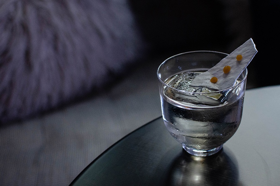 A clear cocktail with a big ice cub is topped with a white rectangular garnish with three jammy orange dots on it.