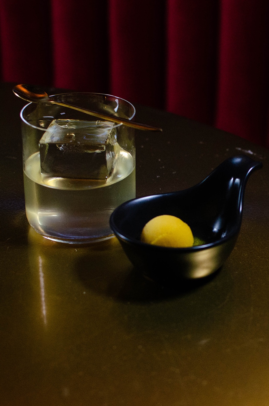 A clear yellow cocktail with a giant ice cube sits next to a black bowl with a small scoop of yellow gelato.