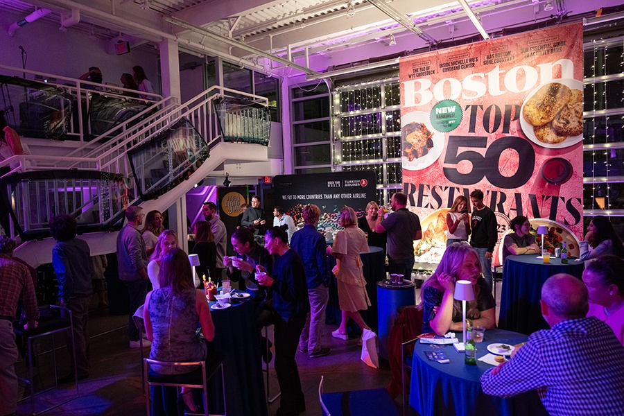 People gather in a dimly lit event space in front of an enlarged copy of Boston magazine's Top 50 Restaurants cover.