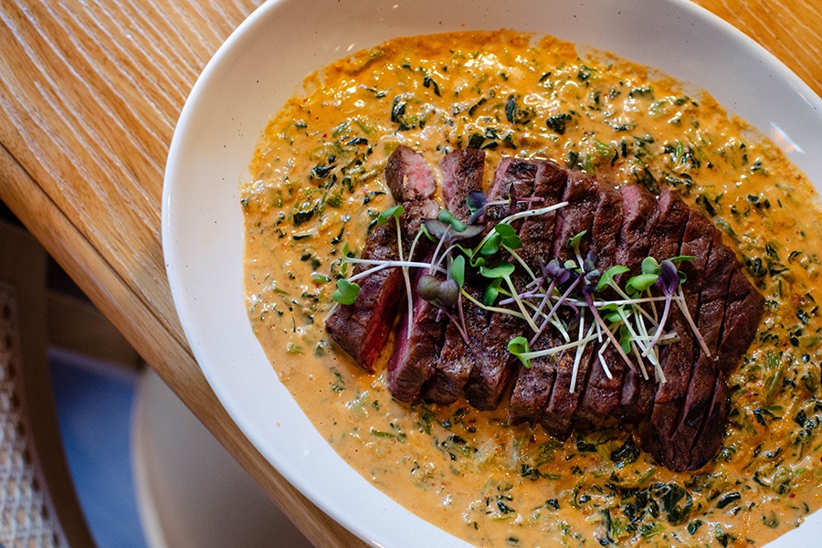 Sliced flat iron steak sits in a pool of thick orange curry full of cooked spinach.