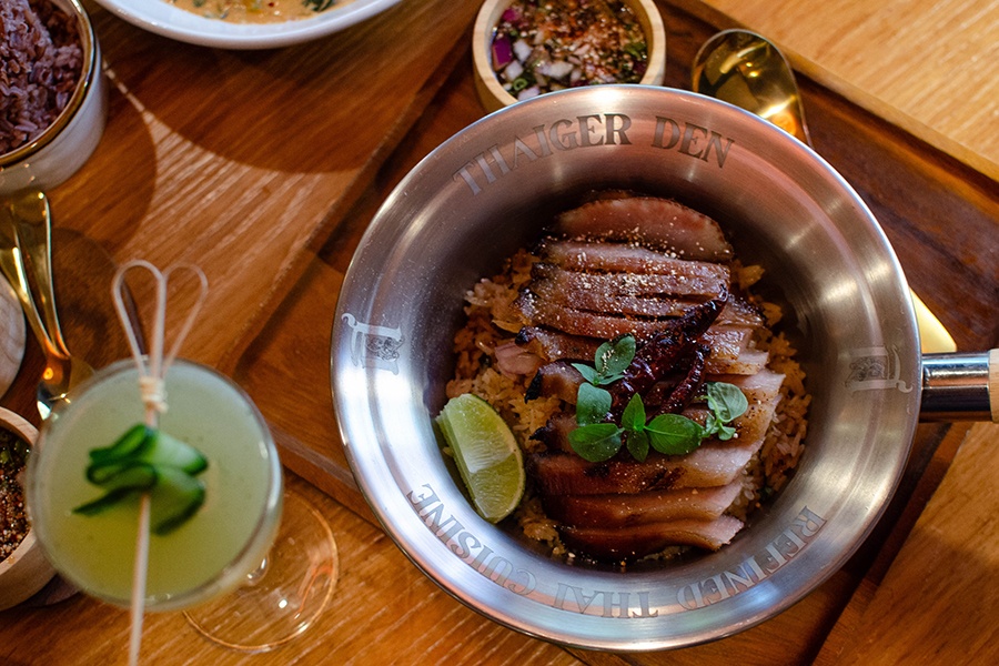 Slices of pork sit atop rice in a silver bowl that says Thaiger Den Refined Thai Cuisine around the edge.