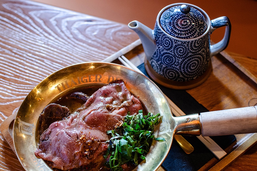 Thinly sliced rare beef sits in broth in a golden bowl, next to a blue and white teapot.