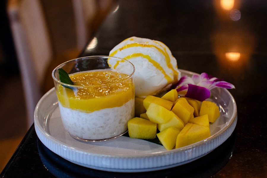 A glass bowl of rice pudding is topped with a mango puree, with sliced mango and coconut ice cream on the side.