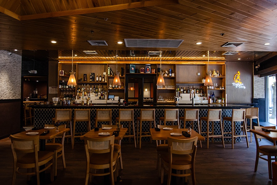 The bar in a restaurant with neon signage reading Thaiger Den, a light wooden ceiling, and rattan-accented chairs.