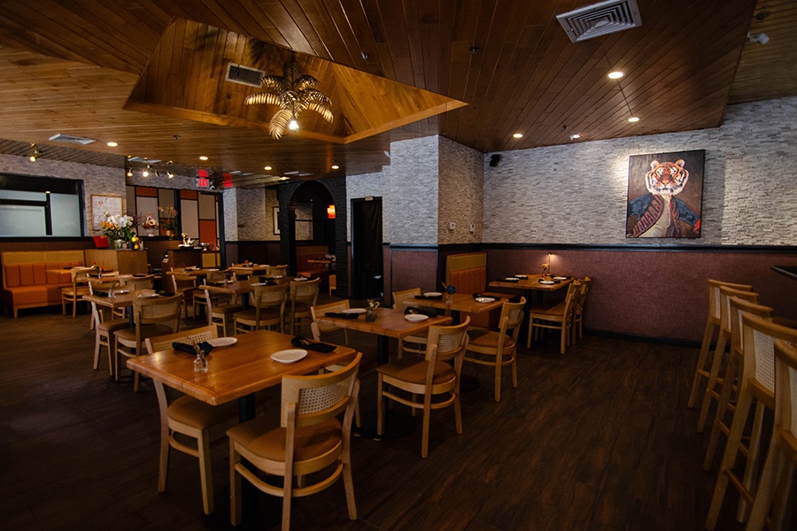 A restaurant interior features a palm-shaped light fixture, a painting of a tiger wearing royal clothes, and light wooden tables with rattan-backed chairs.