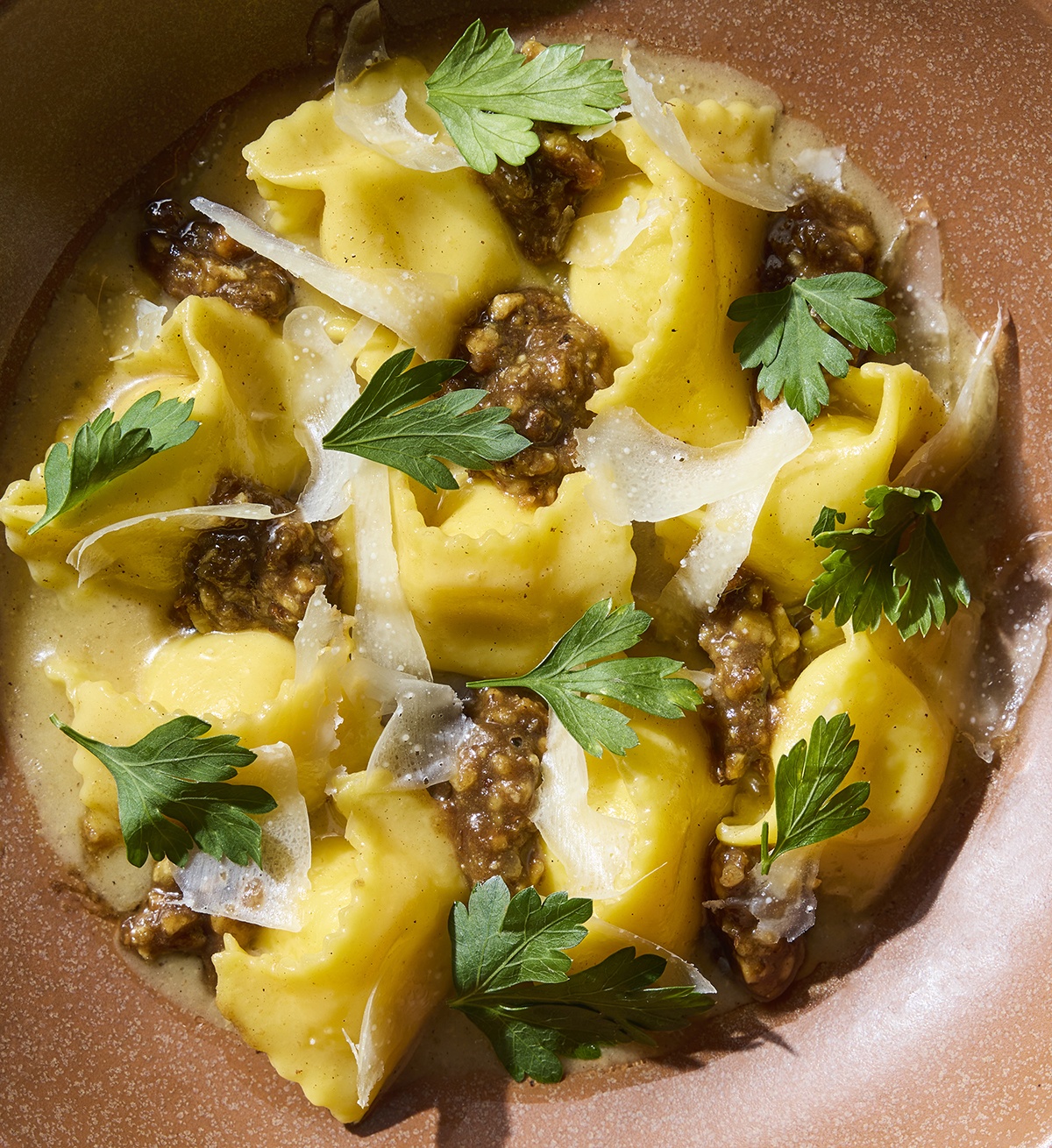 Closeup view on a bowl of pasta, featuring plump agnolotti garnished with thin slivers of cheese and dollops of a brown pesto-like sauce.