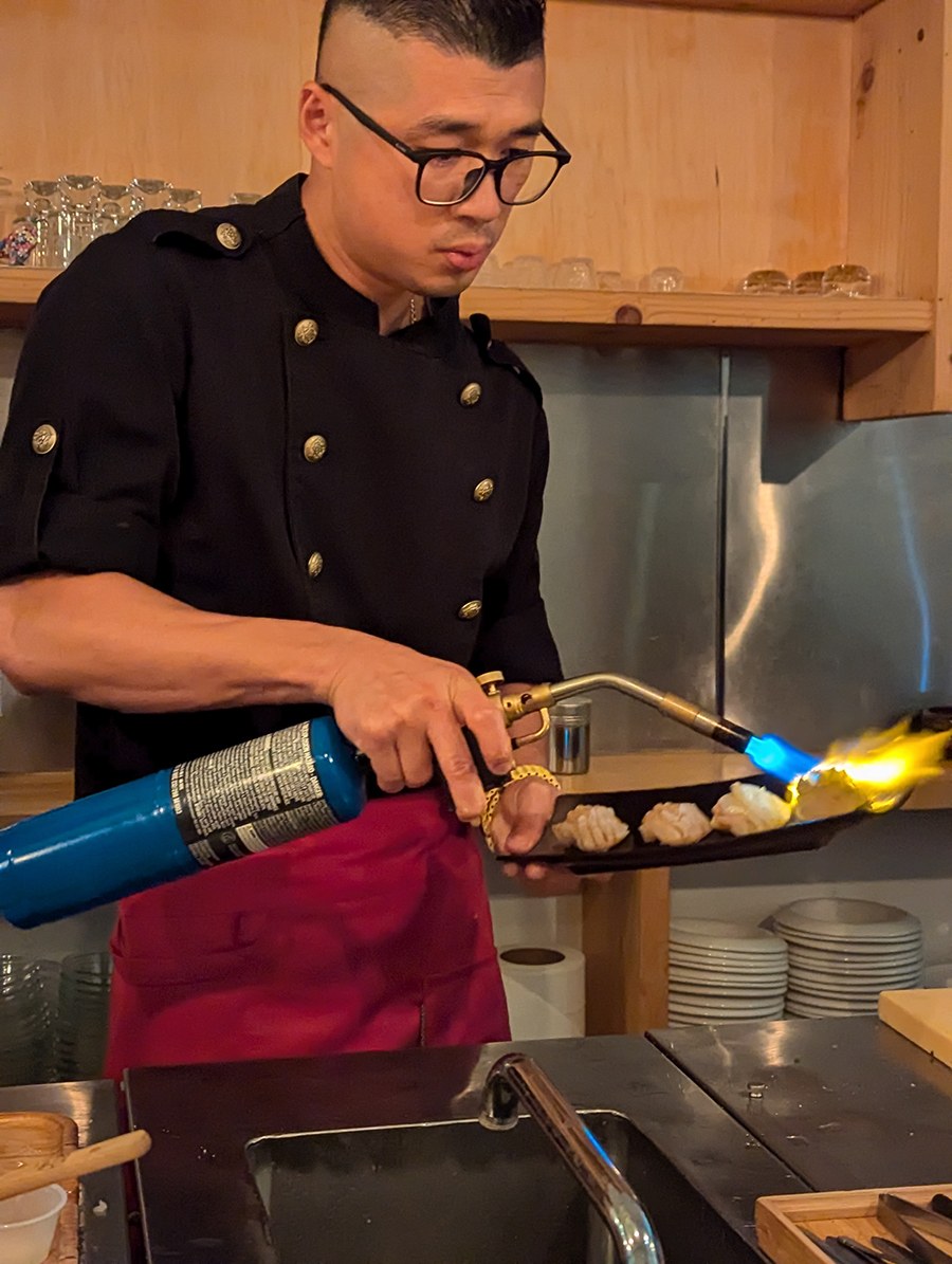 A sushi chef holds a blowtorch to fish behind a sushi bar.