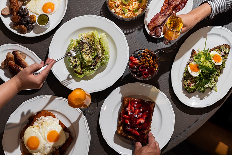 Overhead view of upscale-looking brunch dishes, including bacon, eggs, berry-topped French toast, and Caesar salad.