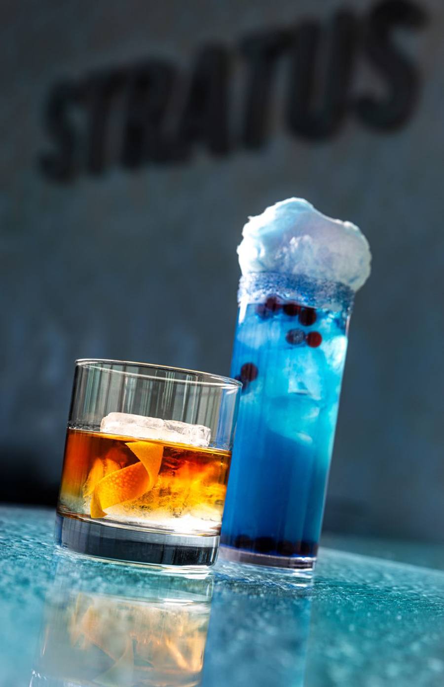 An Old Fashioned-style cocktail and a tall blue cocktail topped with a cloud-like garnish sit on a table that looks like blue ice. The word Stratus is visible on the wall behind.