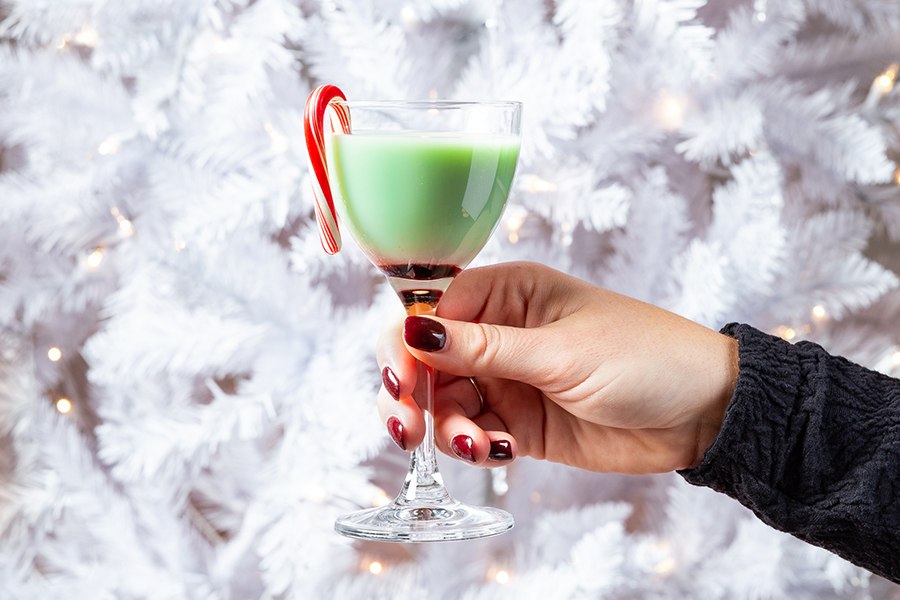 A hand holds a green cocktail with a mini candy cane in front of a white Christmas tree.