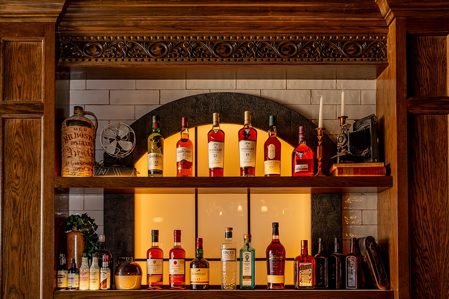 A backbar features intricate woodwork, white bricks, an illuminated false window, and bottles of mostly brown liquors.