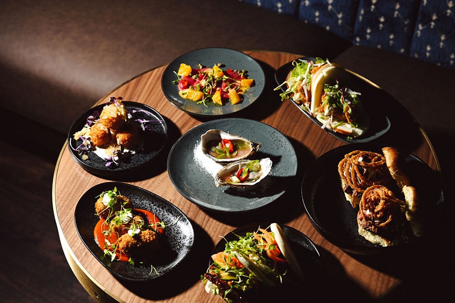 A table of black plates, each topped with elegantly presented bar snacks, including oysters, steak sliders, and more.