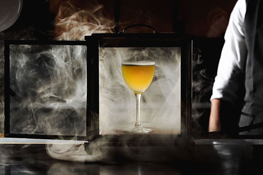 A yellow cocktail is presented in a glass box full of smoke.