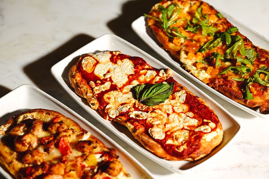 Three oblong flatbreads with various toppings are displayed on white plates on a white marble surface.