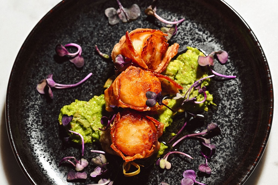 Overhead view of three bacon-wrapped seared scallops sitting atop a guacamole-like puree on a black plate.