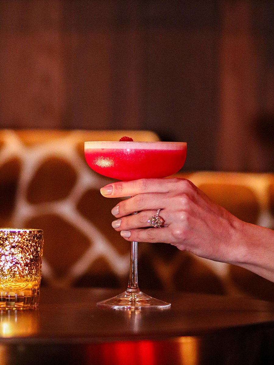 A hand holds a bold pink cocktail in front of a giraffe-print banquette.