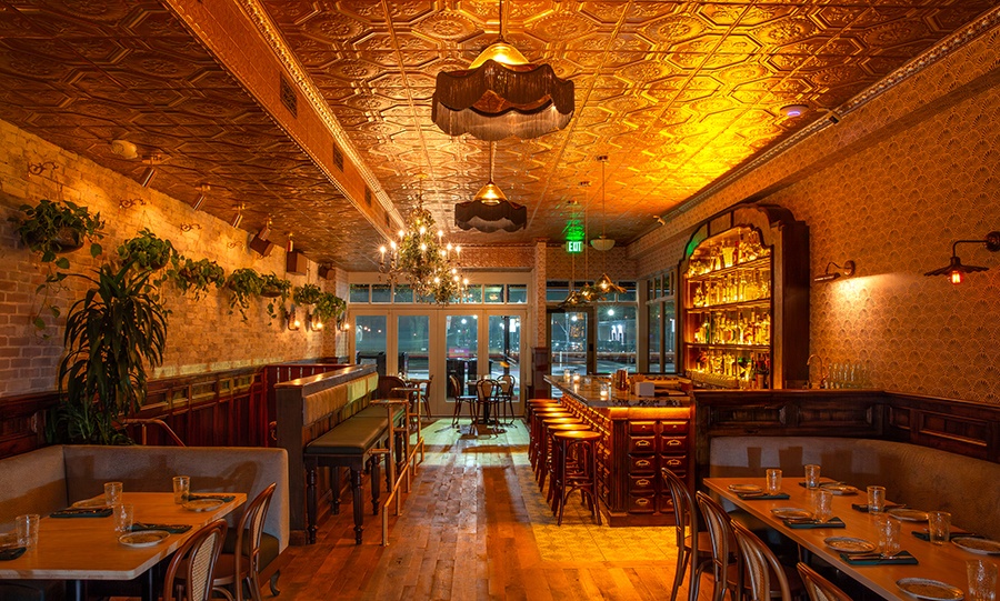 A dimly lit dining room and bar glows golden and features lots of plants.