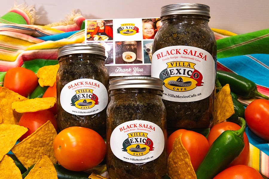 Three jars of a dark salsa with Villa Mexico Cafe labels sit on a colorful background surrounded by chips and chili peppers.