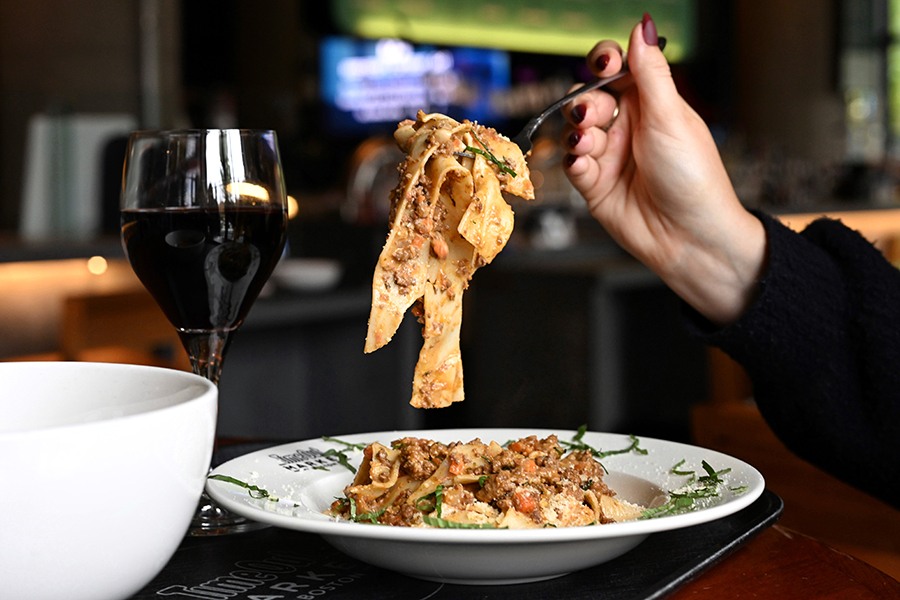 A hand pulls thick noodles topped with a meaty Bolognese sauce off of a plate.