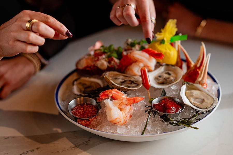 Hands reach toward a seafood platter on a white marble table. There are oysters, shrimp, condiments, and more.