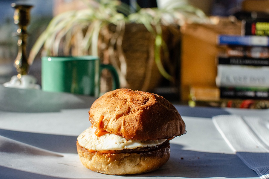 A breakfast sandwich sits on a sunlit counter in front of books and plants.