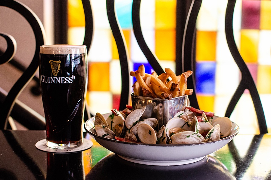 Clams, fries, and a glass of Guinness are displayed on a dark wooden table in front of colorful stained glass.