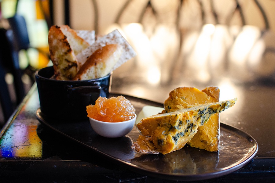 Cheeses, jams, and breads sit on an oval plate on a dark wooden table.