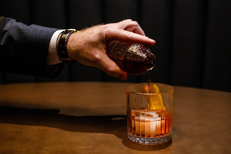 A hand pours a brown cocktail into a glass over a big ice cube.