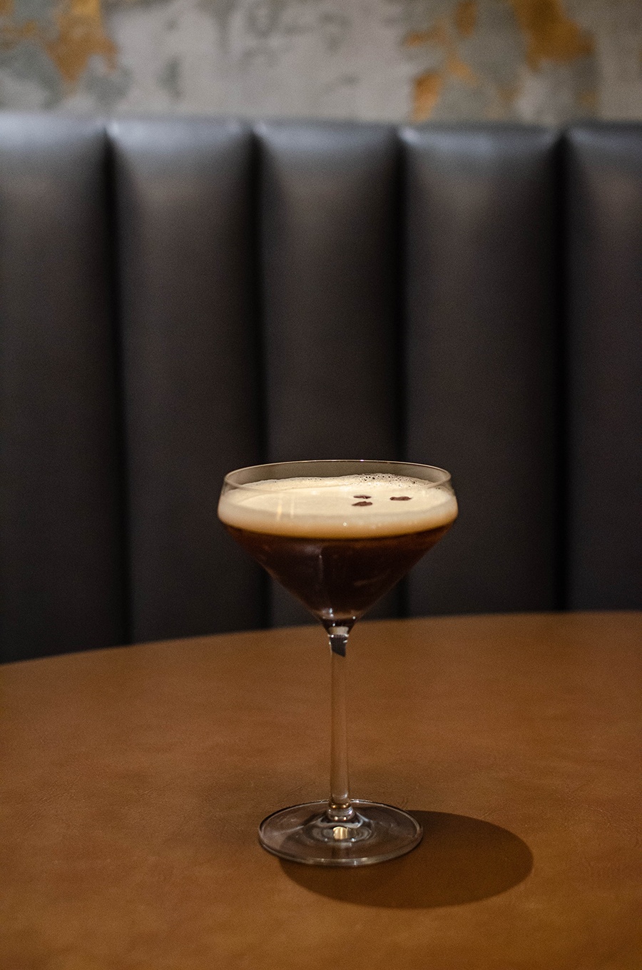 An espresso martini sits on a light brown tabletop with a leather booth in the background.