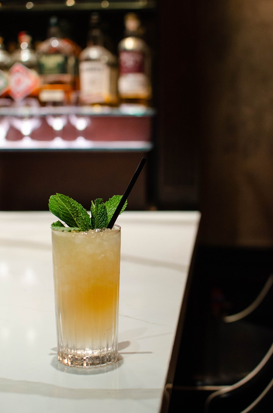 A pale pink-orange cocktail on crushed ice sits on a white marble bar top with bottles of liquor in the background.