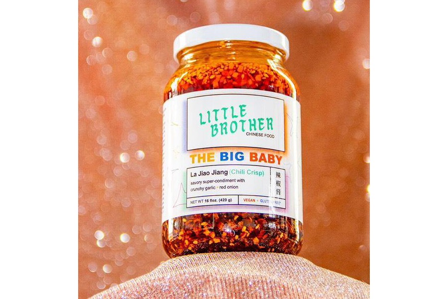 A big bottle of chili crisp with a label from the brand Little Brother is displayed in front of a glittery orange backdrop.