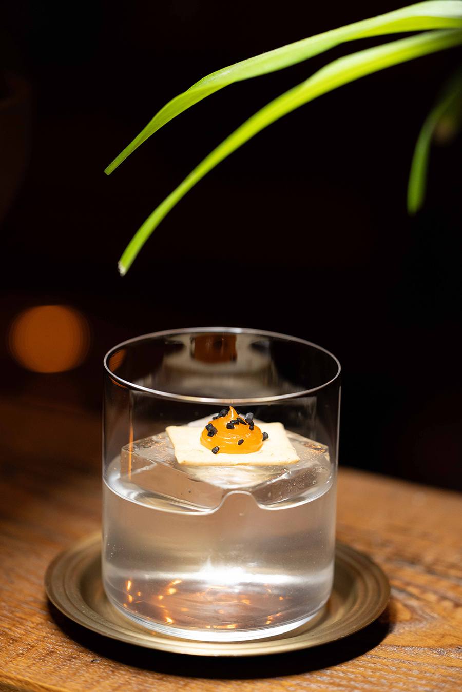 A clear cocktail is garnished with a yellow dollop of gel atop a large ice cube.