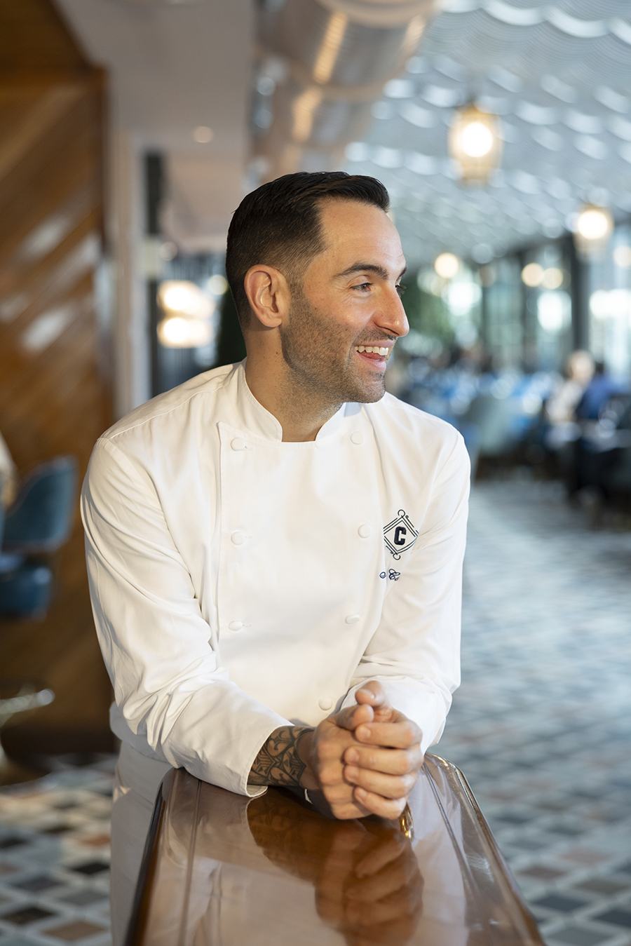 A man in a white chef's coat sits in a restaurant, looking to the side and smiling.