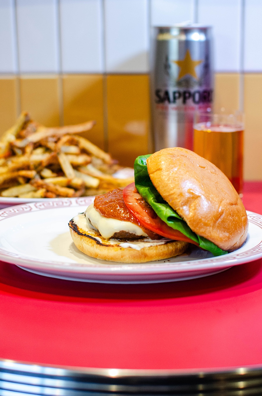 A burger, fries, and Sapporo are displayed on a red table in front of white and yellow tiles.