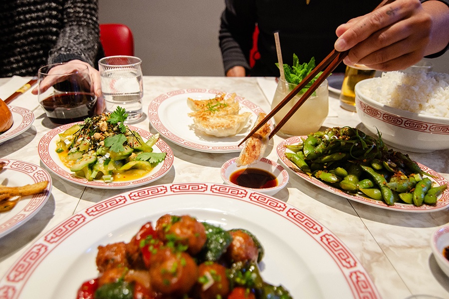 A hand holds chopsticks, dipping a dumpling into sauce on a table spread with food.