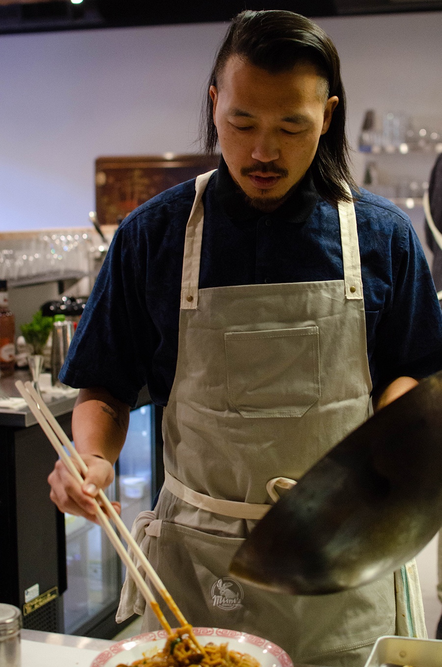 A chef holds a wok in one hand and long chopsticks in the other, adjusting noodles on a plate.